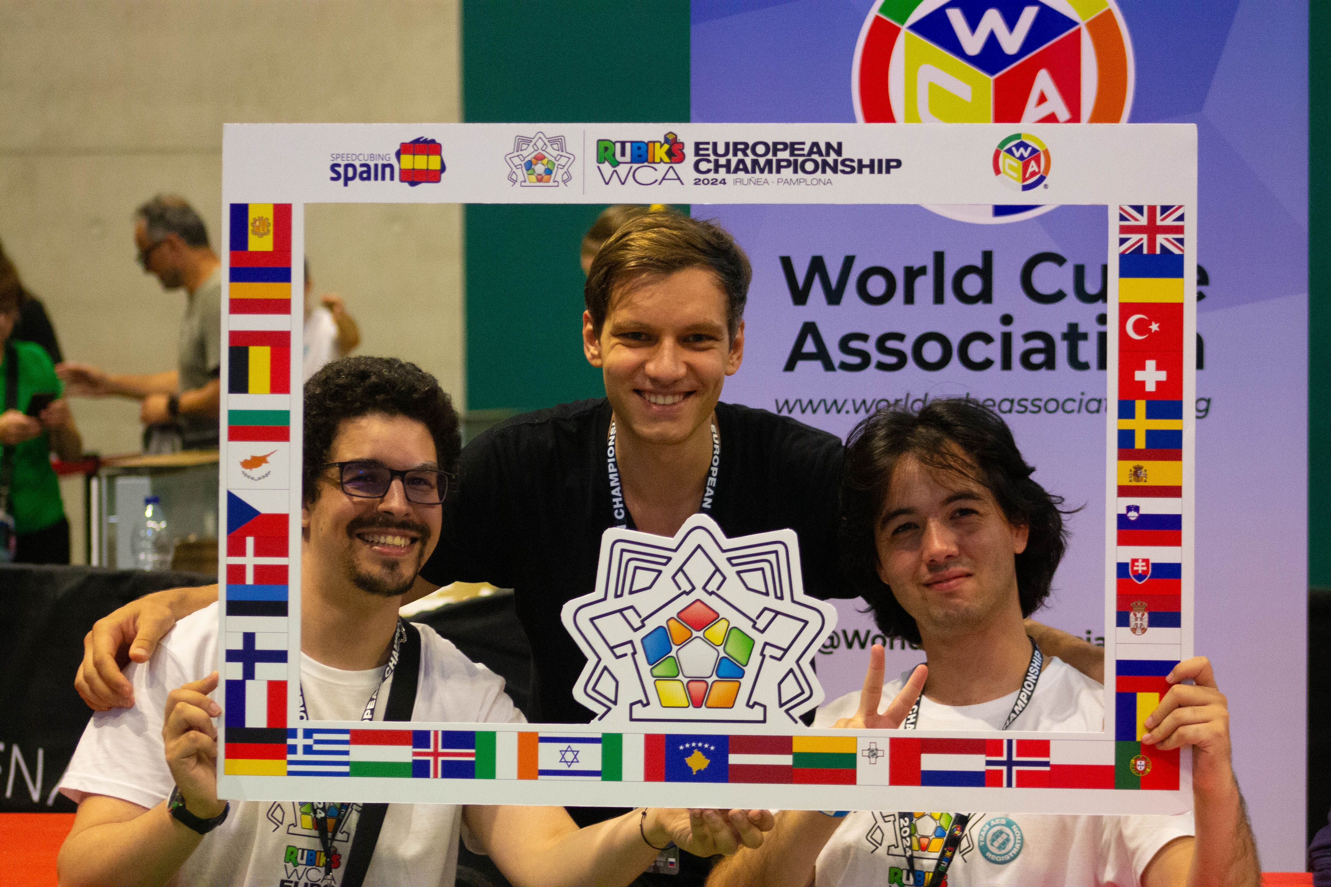 Three speedcubers pose inside a frame decorated with flags and the World Cube Association logo